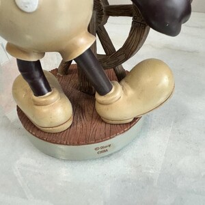 May include: A figurine of a cartoon character in tan and black, holding a wooden ship's wheel. The figurine is standing on a brown base with the text "&copy; Disney CHINA". The character's large shoes are tan.