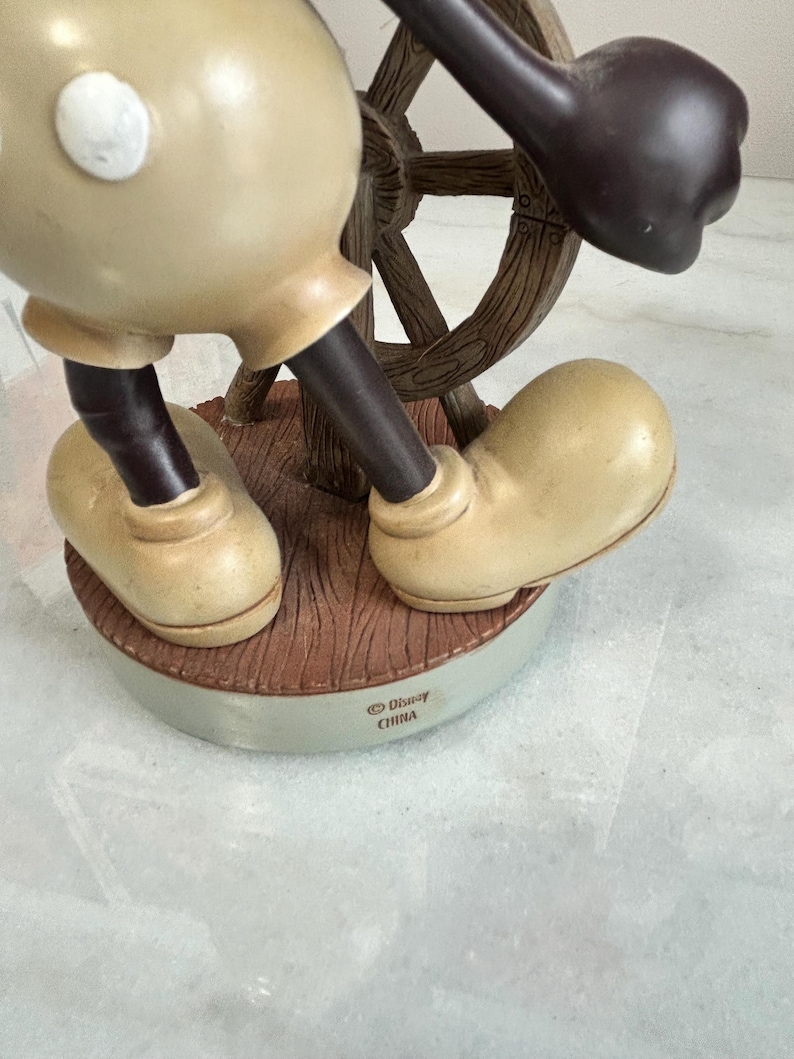 May include: A figurine of a cartoon character in tan and black, holding a wooden ship's wheel. The figurine is standing on a brown base with the text "&copy; Disney CHINA". The character's large shoes are tan.