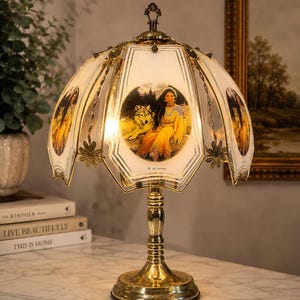 May include: A table lamp with a polished gold-tone metal base and a multi-paneled glass shade. Each glass panel features a circular illustration of a person in traditional attire sitting beside a wolf. The shade is accented with metal floral details.