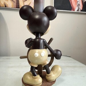 May include: A brown and tan Mickey Mouse figurine in a sailor outfit, holding a ship's wheel. The figurine has large round ears, a chef's hat, and oversized shoes. The base is a light green color with the Disney logo.