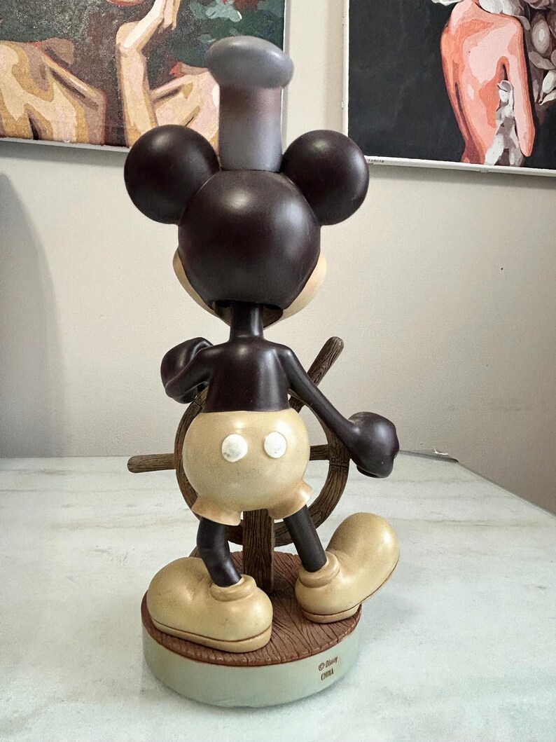 May include: A brown and tan Mickey Mouse figurine in a sailor outfit, holding a ship's wheel. The figurine has large round ears, a chef's hat, and oversized shoes. The base is a light green color with the Disney logo.