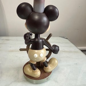 May include: A Mickey Mouse figurine in a chef's hat, steering a ship's wheel. The figure is brown, with tan shorts and shoes. The base is a wooden-look circle. The hat is gray and pink. The figurine is a collectible.
