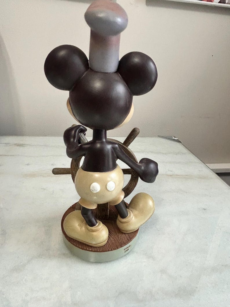 May include: A Mickey Mouse figurine in a chef's hat, steering a ship's wheel. The figure is brown, with tan shorts and shoes. The base is a wooden-look circle. The hat is gray and pink. The figurine is a collectible.