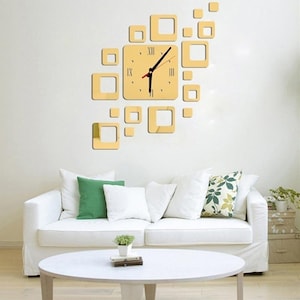 Peut inclure: Une horloge murale moderne avec un cadran carré doré et des aiguilles noires. L'horloge est entourée de cadres carrés dorés de différentes tailles. L'horloge est fixée sur un mur blanc au-dessus d'un canapé blanc avec des coussins verts et blancs.