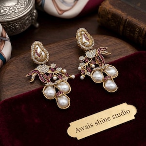 May include: A pair of gold-tone drop earrings featuring white baroque pearls, small clear crystals, and red stones arranged in a floral and vine-like motif. A rectangular gold-colored metal tag with the text Awais shine studio is included.