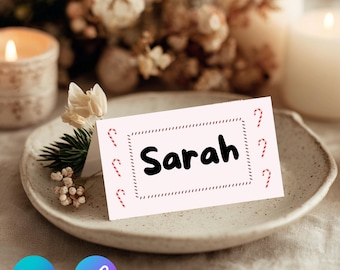 May include: A light pink place card with the name "Sarah" in black, surrounded by a dotted border and candy cane accents. The card sits on a ceramic plate with floral and candle decorations in the background, creating a festive holiday theme.