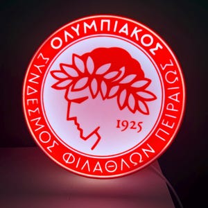 May include: A round, red and white illuminated sign featuring the text "ΣΥΝΔΕΣΜΟΣ ΦΙΛΑΘΛΩΝ ΠΕΙΡΑΙΩΣ ΟΛΥΜΠΙΑΚΟΣ" in Greek lettering. The sign also includes a profile of a person wearing a laurel wreath and the year "1925".