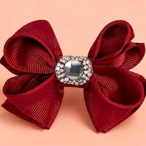May include: A deep burgundy hair bow crafted from ribbed fabric, featuring a central rectangular embellishment composed of a large, clear, faceted centre stone surrounded by a border of small, sparkling clear rhinestones.