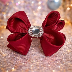 Può includere: Un fiocco rosso acceso con un centro di strass quadrato e scintillante. Il fiocco è realizzato in tessuto a coste e lo sfondo ha un effetto bokeh scintillante.