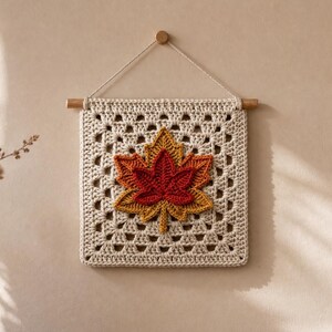 Peut inclure: Une tenture murale carrée en crochet composée d'une base granny square couleur crème avec un motif de feuille d'érable superposé en trois dimensions dans des tons rouge foncé, orange et jaune, suspendue à une tige en bois avec une cordelette.