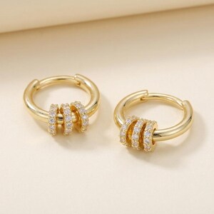 May include: A pair of gold hoop earrings, each featuring three small, circular charms encrusted with clear, sparkling stones. The hoops have a polished, smooth finish and a hinged closure mechanism, creating a cohesive and reflective appearance.