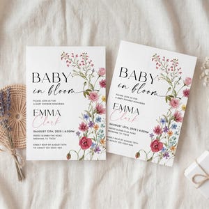 May include: Two white baby shower invitations with floral designs and the text "Baby in Bloom." The invitations are adorned with watercolor flowers in pink, purple, and blue. A small gift box and lavender bundle are also present.