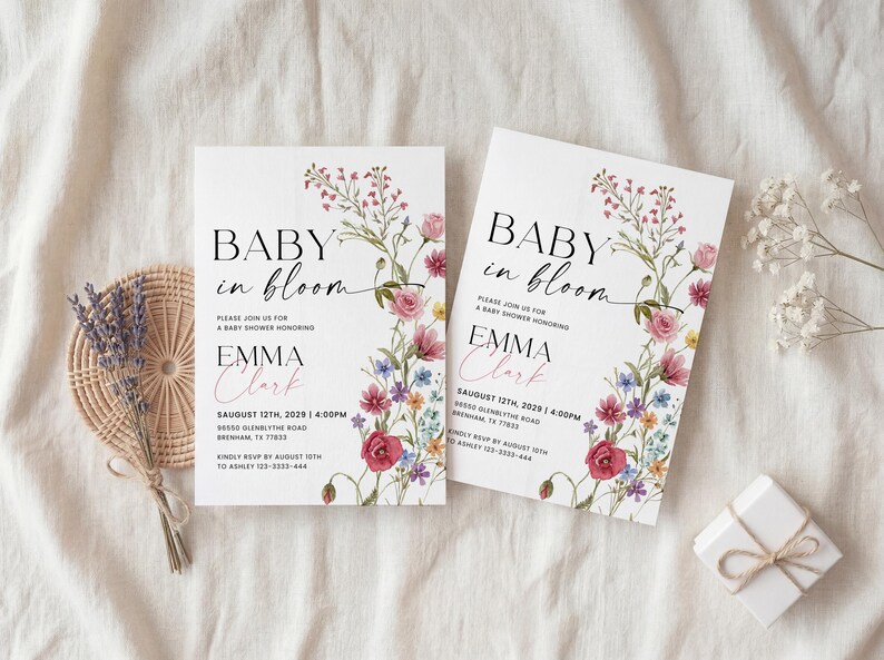 May include: Two white baby shower invitations with floral designs and the text "Baby in Bloom." The invitations are adorned with watercolor flowers in pink, purple, and blue. A small gift box and lavender bundle are also present.
