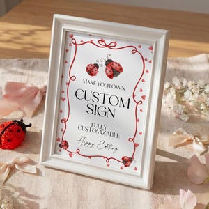 May include: A white framed sign with the text "MAKE YOUR OWN CUSTOM SIGN" and "FULLY CUSTOMIZABLE Happy Eating". The sign is decorated with red ladybugs and hearts. A red ladybug figurine and pink bows are in the foreground.