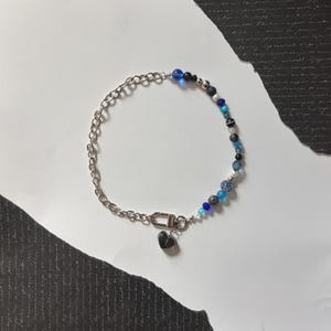 May include: A silver chain necklace with a mix of blue, black, and clear beads. A small black heart charm hangs from the clasp. The beads vary in size and shape, creating a unique and stylish accessory.