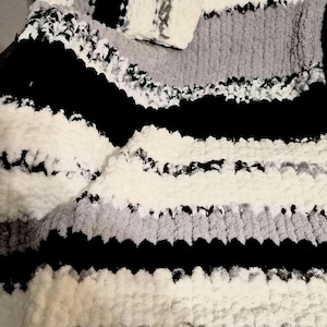 Könnte beinhalten: Eine weiche, grob gestrickte Decke mit Streifenmuster. Die Decke hat abwechselnd weiße, graue und schwarze Streifen. Die Textur wirkt flauschig und gemütlich, geeignet für die Inneneinrichtung.