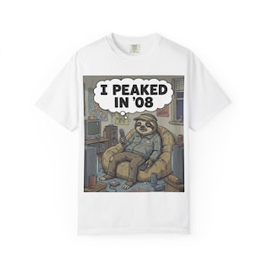 May include: White t-shirt featuring a cartoon sloth lounging in a beanbag chair, holding a remote control. The sloth is wearing a hat and a button-down shirt. A thought bubble above the sloth reads, "I peaked in '08."