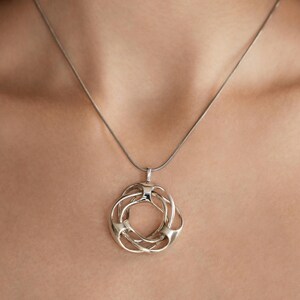 Op de afbeelding: Een zilverkleurige hangerketting met een ingewikkeld, open ontwerp. De hanger bestaat uit onderling verbonden lussen en rondingen, die aan een fijne ketting hangen. De ketting wordt gedragen op een neutrale huidskleur.