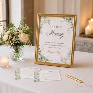 May include: A gold framed sign with the text "Share a Memory" and floral accents. Beside it are matching cards and a gold pen. A vase of white roses and a lit candle are also on the table, creating a memorial or event theme.