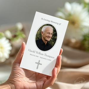 May include: A white memorial card held in a hand, featuring a photo of a smiling older man in a black shirt. The card reads "In Loving Memory of Harold William Thompson" with dates and a silver cross graphic.