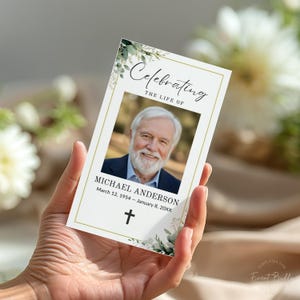 May include: A memorial card held in a hand, featuring a portrait of a smiling older man. The card has a gold border and includes the text "Celebrating The Life Of" and the name "MICHAEL ANDERSON" with dates. Greenery and a cross are also featured.