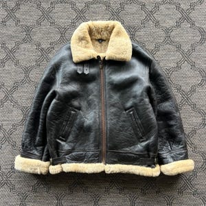 Puede incluir: Una chaqueta de cuero marrón oscuro con un cuello grueso de borrego beige y puños a juego. La chaqueta incluye una cremallera central de metal, dos bolsillos verticales y correas laterales ajustables en el dobladillo para un ajuste seguro.