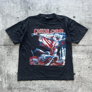 Puede incluir: Una camiseta negra de manga corta con un estampado gráfico que incluye el texto Cannibal Corpse en una fuente roja que gotea. La ilustración muestra una figura esquelética en una cruz sobre una figura tendida, rodeada de velas.