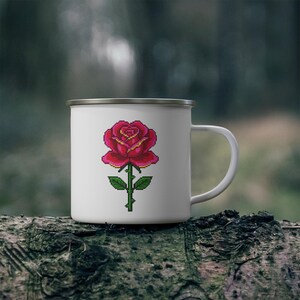 May include: White enamel mug with a pixelated red rose design. The rose has yellow accents and a green stem with leaves. The mug has a silver rim and a white handle. The mug is sitting on a log.