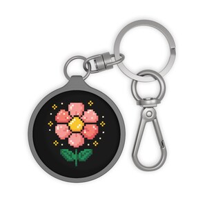 May include: A round, black keychain with a pixelated flower design. The flower has pink petals, a yellow center, and green leaves. The keychain has a silver clasp and a key ring.