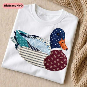 Peut inclure: T-shirt blanc avec un motif de canard en patchwork. Le canard a une tête bleu marine avec des étoiles blanches, un bec orange et un corps bordeaux avec un motif cachemire. Les ailes et le corps sont faits de divers tissus à motifs.