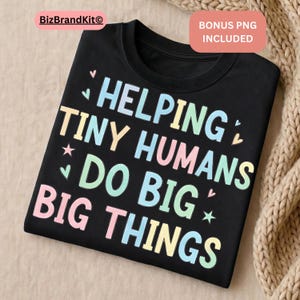 May include: Black t-shirt with the text "HELPING TINY HUMANS DO BIG THINGS" in pastel colors. The design includes hearts and stars. The shirt is folded and displayed on a neutral surface.