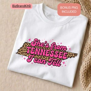 Puede incluir: Camiseta blanca con un diseño rosa y estampado de leopardo. El diseño presenta el texto "She's from TENNESSEE I can Tell" en una fuente retro, con un contorno de leopardo del estado de Tennessee.
