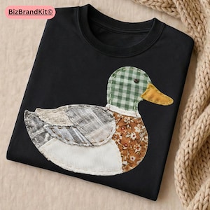 May include: Black crew neck t-shirt with a patchwork duck design. The duck has a green and white checked head, a yellow beak, and a body made of various patterned fabrics in brown, beige, and white. The design is centered on the shirt.