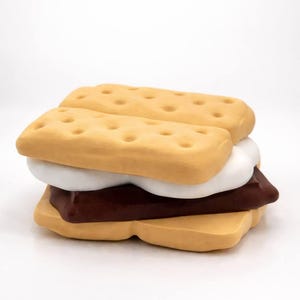 May include: A whimsical, oversized s'more featuring two tan graham crackers with dot details, a white marshmallow, and a dark brown chocolate bar. The dessert is stacked on a white background.