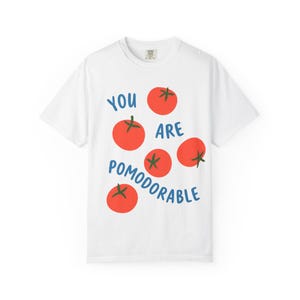 May include: White t-shirt with a playful design featuring red tomatoes and the blue text "YOU ARE POMODORABLE". The shirt has a crew neck and short sleeves, perfect for casual wear.