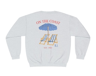 May include: Light gray sweatshirt with a beach-themed graphic. The design features a blue umbrella, two beach chairs with blue and white stripes, and the text "ON THE COAST" in red. Below the graphic is "EST. 1985" and "K.I."