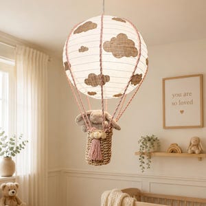 May include: A hot air balloon pendant light featuring a white paper lantern shade with brown cloud patterns, suspended by pink twisted ropes. A woven basket hangs below, containing a plush grey rabbit toy with long ears and a pink tassel accent.