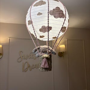 Puede incluir: Lámpara decorativa con forma de globo aerostático con pantalla de papel blanco adornada con motivos de nubes marrones. Un conejito de peluche gris está sentado en una cesta tejida. La lámpara tiene detalles de cuerda y borlas rosas.