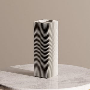Op de afbeelding: Een hoge, gebroken witte vaas met een uniek, getextureerd ontwerp. De vaas heeft een rechthoekige vorm met verticale, golfachtige ribbels die een gestreept patroon creëren. De bovenste opening is cirkelvormig. De vaas is waarschijnlijk van keramiek of een vergelijkbaar materiaal.