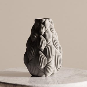 May include: A matte light gray vase with a textured exterior featuring a repeating pattern of vertical, leaf-like or petal-shaped ridges that wrap around the entire body of the vessel, creating a dimensional and organic appearance.
