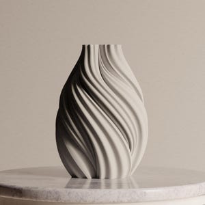 Könnte beinhalten: Eine cremefarbene Vase mit einem einzigartigen, wirbelnden Design. Die Vase hat einen schmalen Hals und einen breiteren Boden, mit vertikalen, wellenartigen Rippen, die eine strukturierte Oberfläche bilden. Sie steht auf einer marmorähnlichen Oberfläche und zeigt ihre elegante Form.