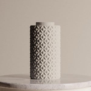Op de afbeelding: Een cilindervormige, crèmekleurige vaas met een geometrisch patroon. De vaas heeft een getextureerd oppervlak en een kleine rand aan de bovenkant. Het staat op een marmeren oppervlak tegen een neutrale achtergrond.
