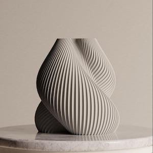 Op de afbeelding: Een crèmekleurige, abstracte vaas met een gedraaid ontwerp. De vaas heeft verticale, geribbelde lijnen die om de vorm wikkelen. Hij staat op een marmeren oppervlak met een neutrale achtergrond.