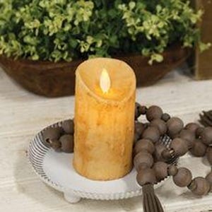 May include: A battery-operated, tan-colored pillar candle with a flickering LED flame sits on a white, scalloped-edge tray. A strand of brown wooden beads with a tassel is draped nearby. A wooden bowl with greenery is in the background.