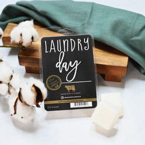 May include: A black wax melt package with the words "LAUNDRY day" in white script. The package is on a wooden surface with cotton stems and a green cloth. The wax melts are 6.6 ounces.