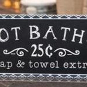 May include: A rectangular black sign with white text that reads "HOT BATHS 25¢ soap & towel extra." The sign has a decorative white border with a geometric pattern. The sign is likely for a bathroom or spa.