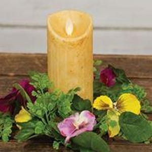 May include: A battery-operated, yellow pillar candle with a flickering LED flame. The candle is surrounded by a wreath of green leaves, purple and yellow flowers, and sits on a wooden surface. The candle is approximately 6 inches tall.