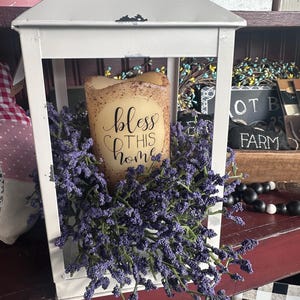 May include: A white metal lantern with a candle that reads "bless this home." The candle is surrounded by purple flowers. The lantern sits on a red shelf, with other decorative items in the background.