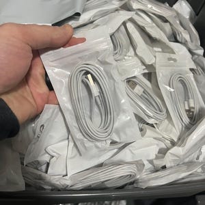Könnte beinhalten: Ein Behälter voller weißer Ladekabel, die jeweils ordentlich aufgerollt und in durchsichtigen Plastiktüten verpackt sind. Die Kabel haben einen USB-Anschluss und einen Lightning-Anschluss. Das Bild zeigt eine große Menge dieser elektronischen Zubehörteile.