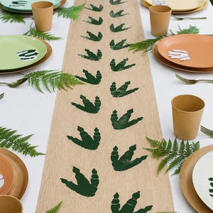 Puede incluir: Un camino de mesa de tono madera clara con un patrón repetitivo de huellas de dinosaurio de color verde oscuro dispuestas en dos filas paralelas, centrado sobre un mantel blanco con platos, vasos y hojas de helecho verdes.
